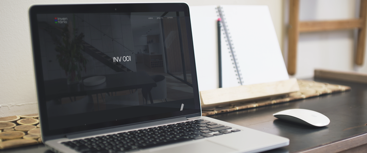 A página inicial do site Inventário aberta em um MacBook, em cima de uma mesa escura, com um mouse branco sem fio à sua direita.