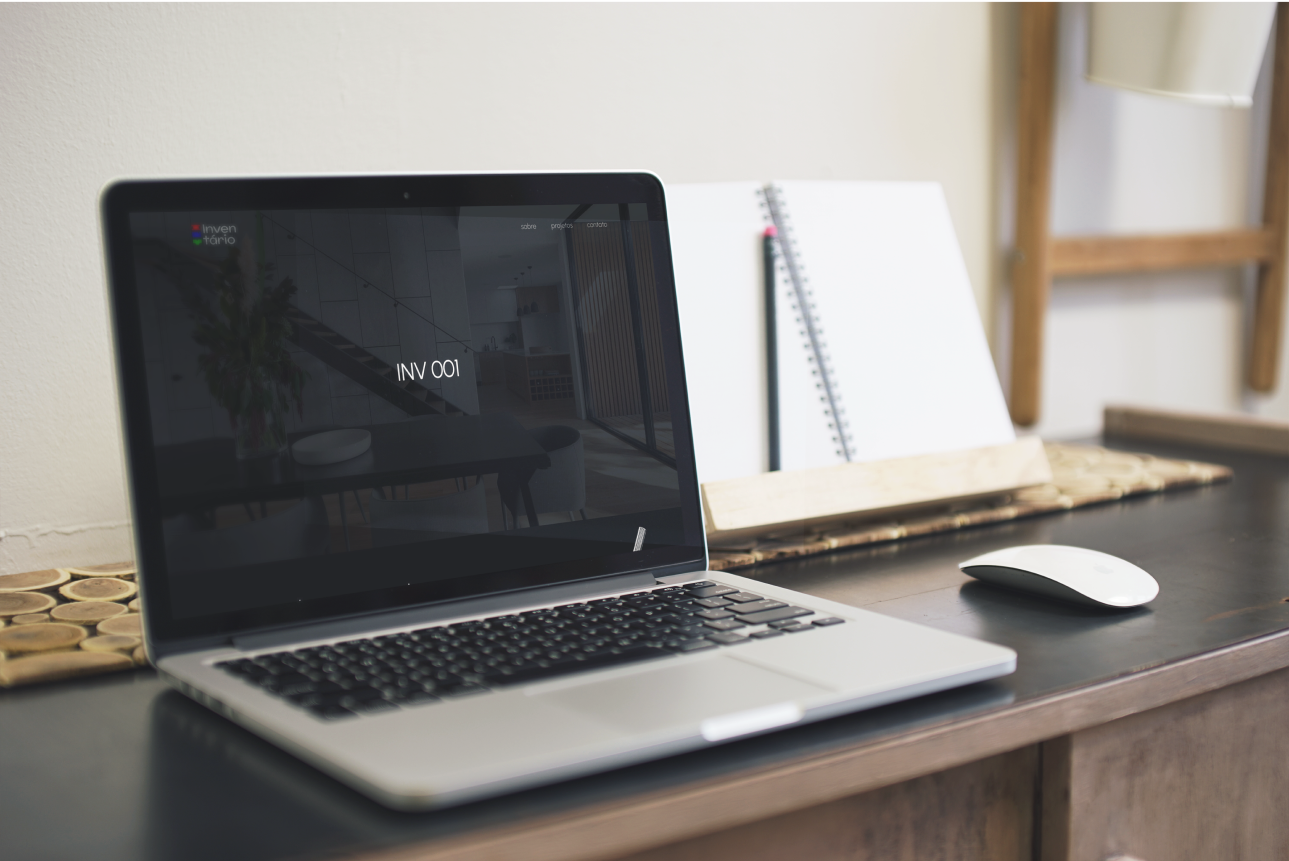 A página inicial do site Inventário aberta em um MacBook, em cima de uma mesa escura, com um mouse branco sem fio à sua direita.
